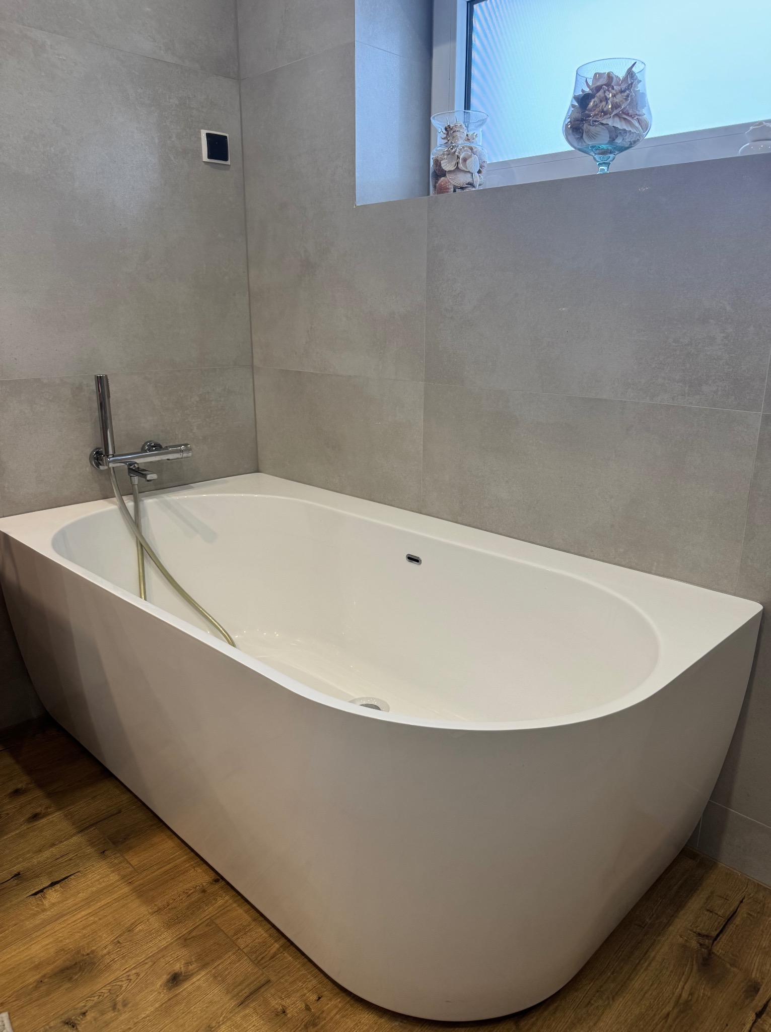 Nowoczesna, biała wanna wolnostojąca w łazience wykończonej szarymi płytkami. Na parapecie okna dekoracje z muszelek. Drewniana podłoga dodaje ciepła minimalistycznemu wnętrzu.