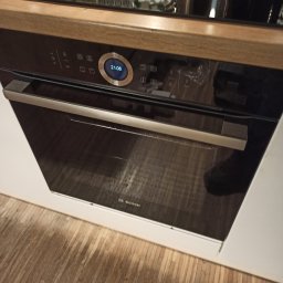 Mazurczak Elektroinstalacje - Nowoczesny piekarnik elektryczny marki Bosch w zabudowie kuchennej, z widocznym panelem sterowania i odbiciem w szklanych drzwiach. Ustawiony na drewnianej podłodze.