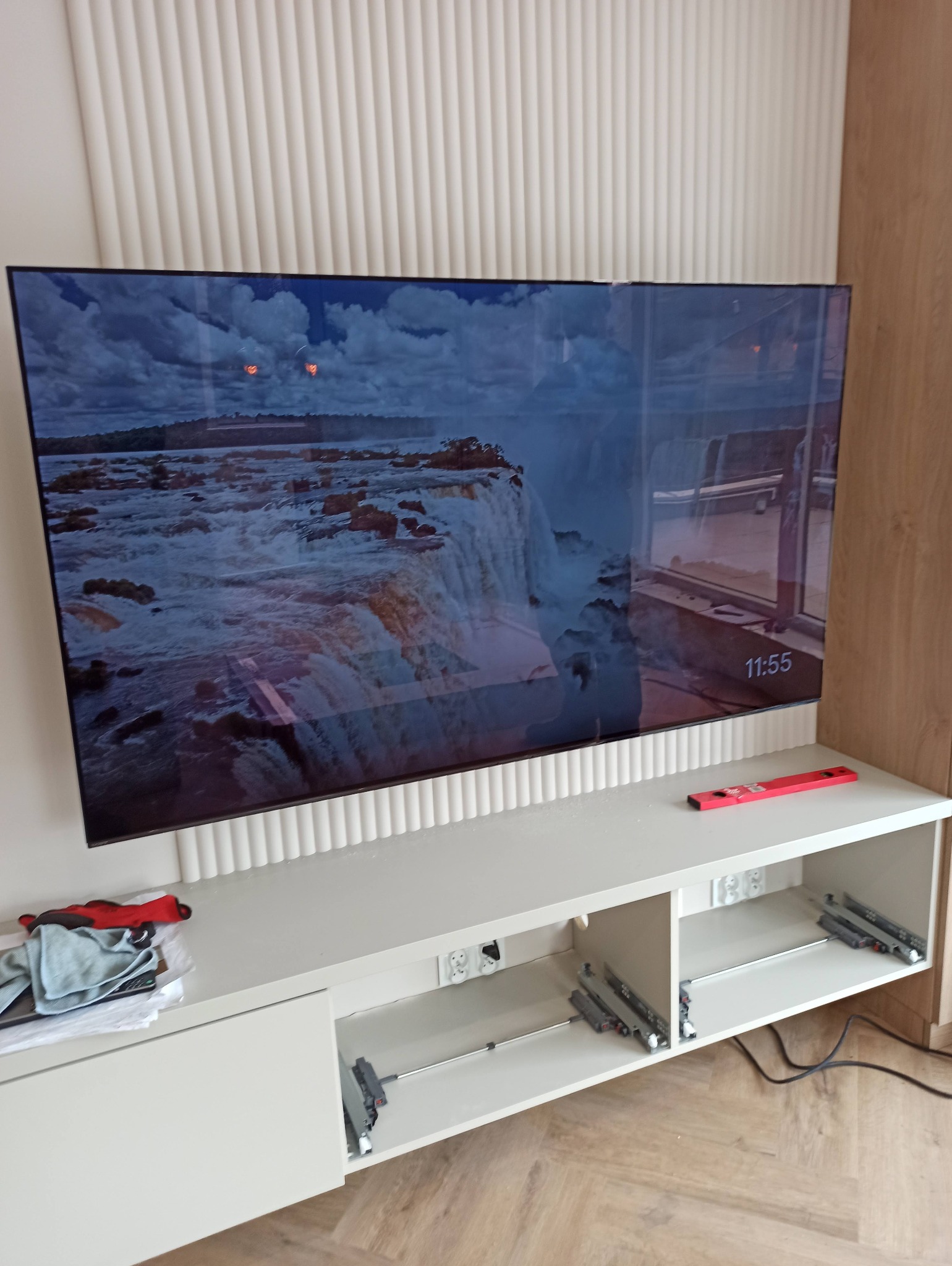 Nowoczesny telewizor LED zawieszony na ścianie z dekoracyjną listwą. Widoczne gniazdka elektryczne i poziomica, sugerujące montaż. Ekran wyświetla krajobraz.