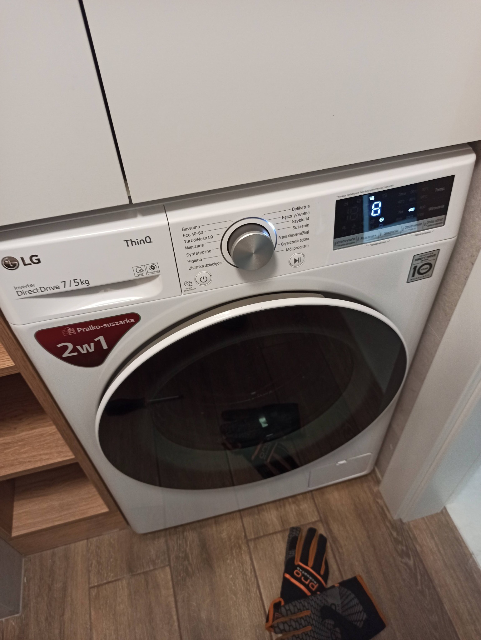 Nowoczesna pralko-suszarka LG w zabudowie kuchennej, widoczne detale panelu sterowania i oznaczenia modelu. Na podłodze leżą rękawice robocze.