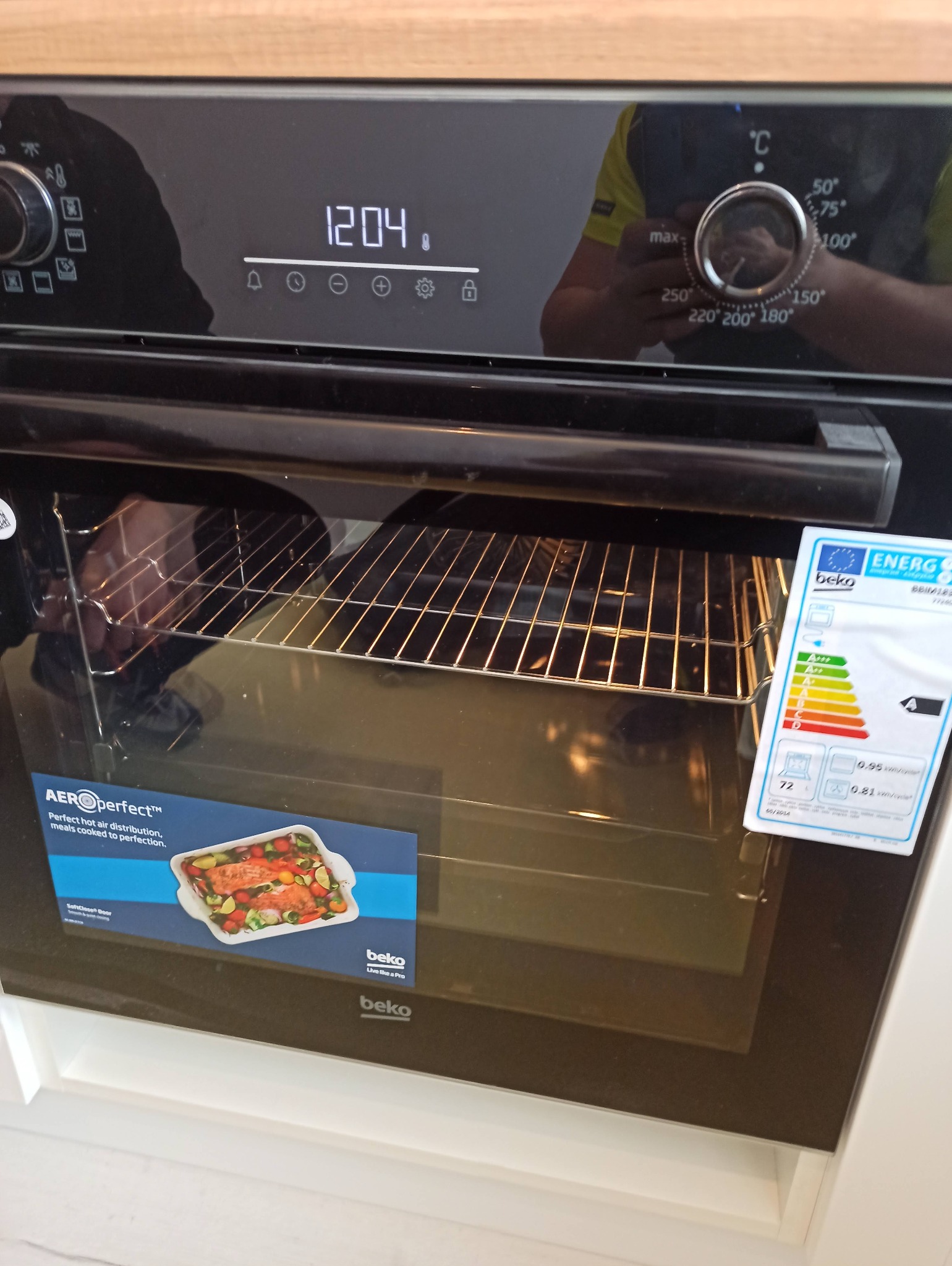 Nowoczesny piekarnik Beko z wyświetlaczem czasu i naklejką energetyczną, widoczna kratka wewnątrz. Odbicie osoby w szklanych drzwiach. Jasne, neutralne tło.