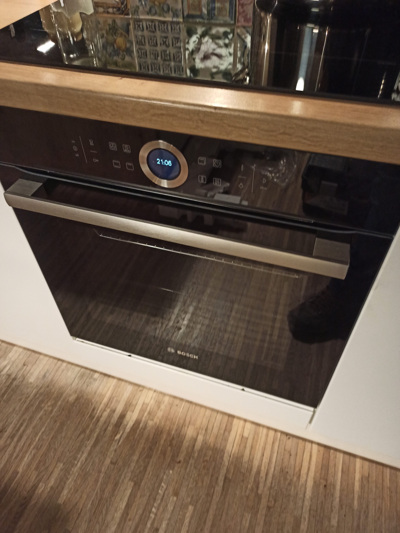 Nowoczesny piekarnik elektryczny marki Bosch w zabudowie kuchennej, z widocznym panelem sterowania i odbiciem w szklanych drzwiach. Ustawiony na drewnianej podłodze.