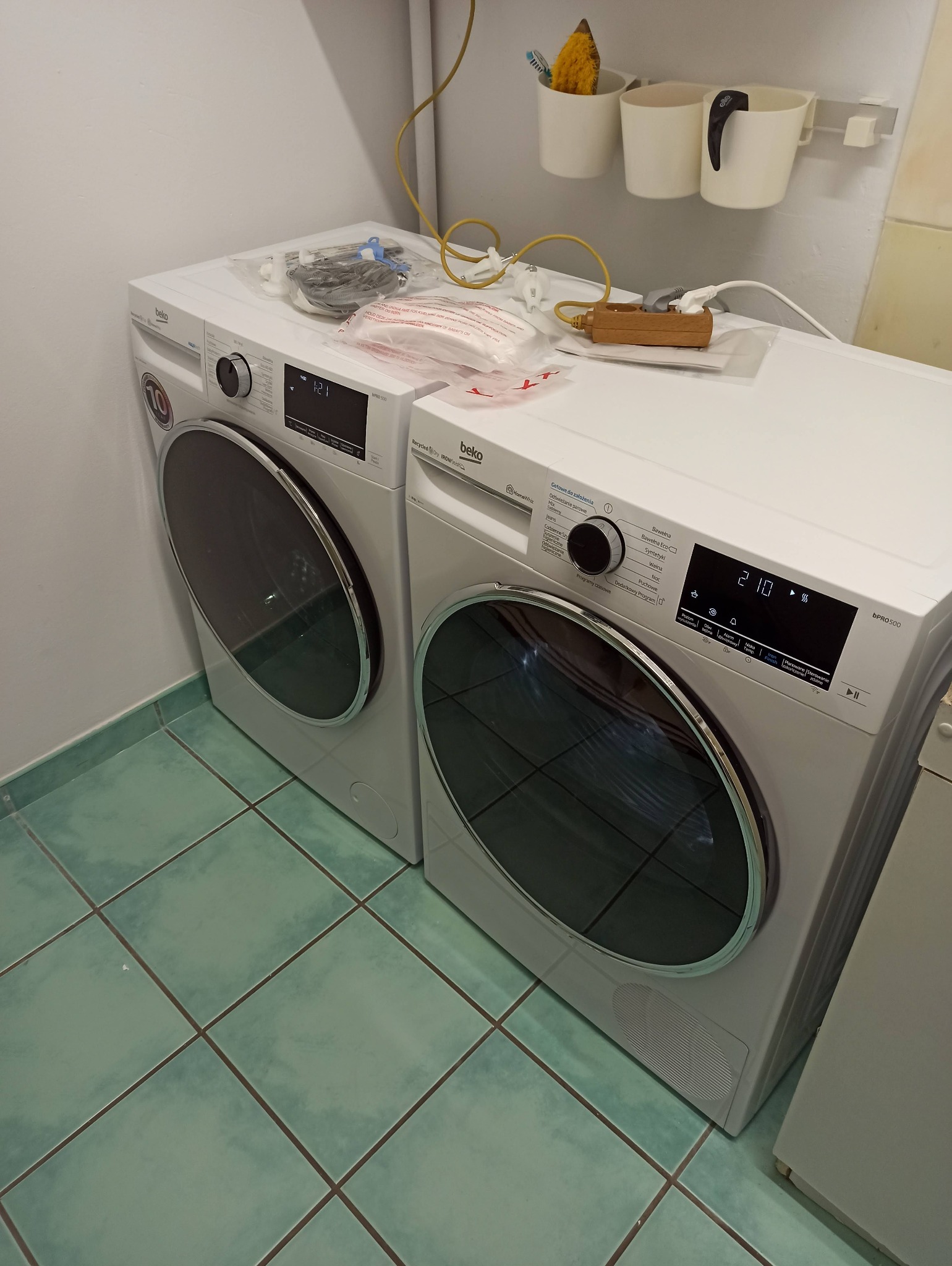 Dwie białe pralki Beko, jedna na drugiej, z akcesoriami i przedłużaczem na górze. Widoczne płytki podłogowe w odcieniach zieleni i białe ściany. Ujęcie z bliska.