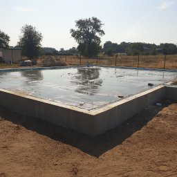 Wylane fundamenty domu jednorodzinnego, widoczne zbrojenia i izolacja, teren budowy z piaskiem i drzewami w tle, słoneczny dzień.