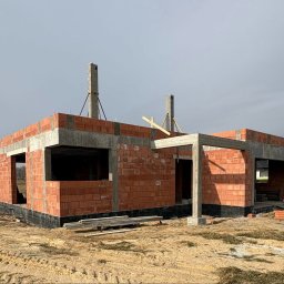 Surowy stan domu z czerwonej cegły, widoczne betonowe nadproża i słupy, teren budowy z piaskiem i resztkami materiałów. Budowa domu w trakcie realizacji.