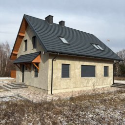 Nowoczesny dom jednorodzinny z szarą dachówką i beżową elewacją. Widoczne okna z roletami zewnętrznymi i drewniane elementy wykończeniowe nad wejściem.