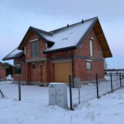 Niedokończony, dwukondygnacyjny dom z czerwonej cegły w zimowej scenerii. Widoczne okna z drewnianymi ramami i czarny dach pokryty śniegiem. Teren ogrodzony siatką.
