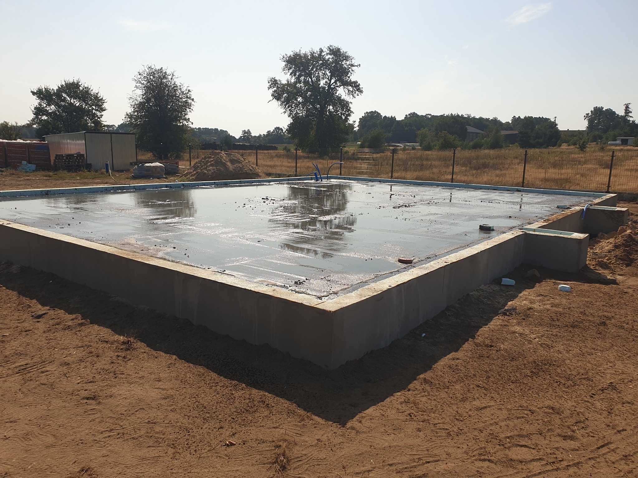 Wylane fundamenty domu jednorodzinnego, widoczne zbrojenia i izolacja, teren budowy z piaskiem i drzewami w tle, słoneczny dzień.