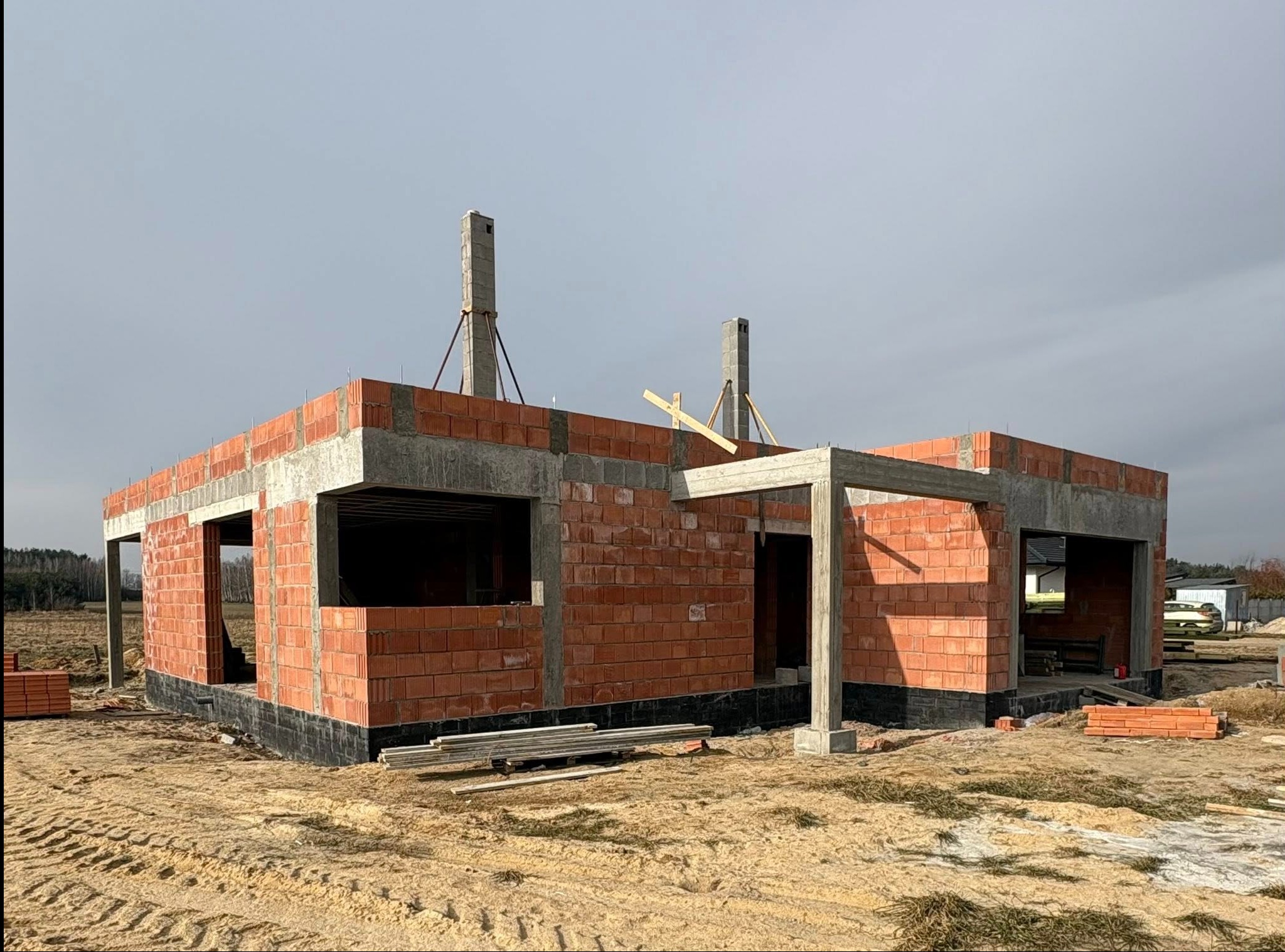 Surowy stan domu z czerwonej cegły, widoczne betonowe nadproża i słupy, teren budowy z piaskiem i resztkami materiałów. Budowa domu w trakcie realizacji.