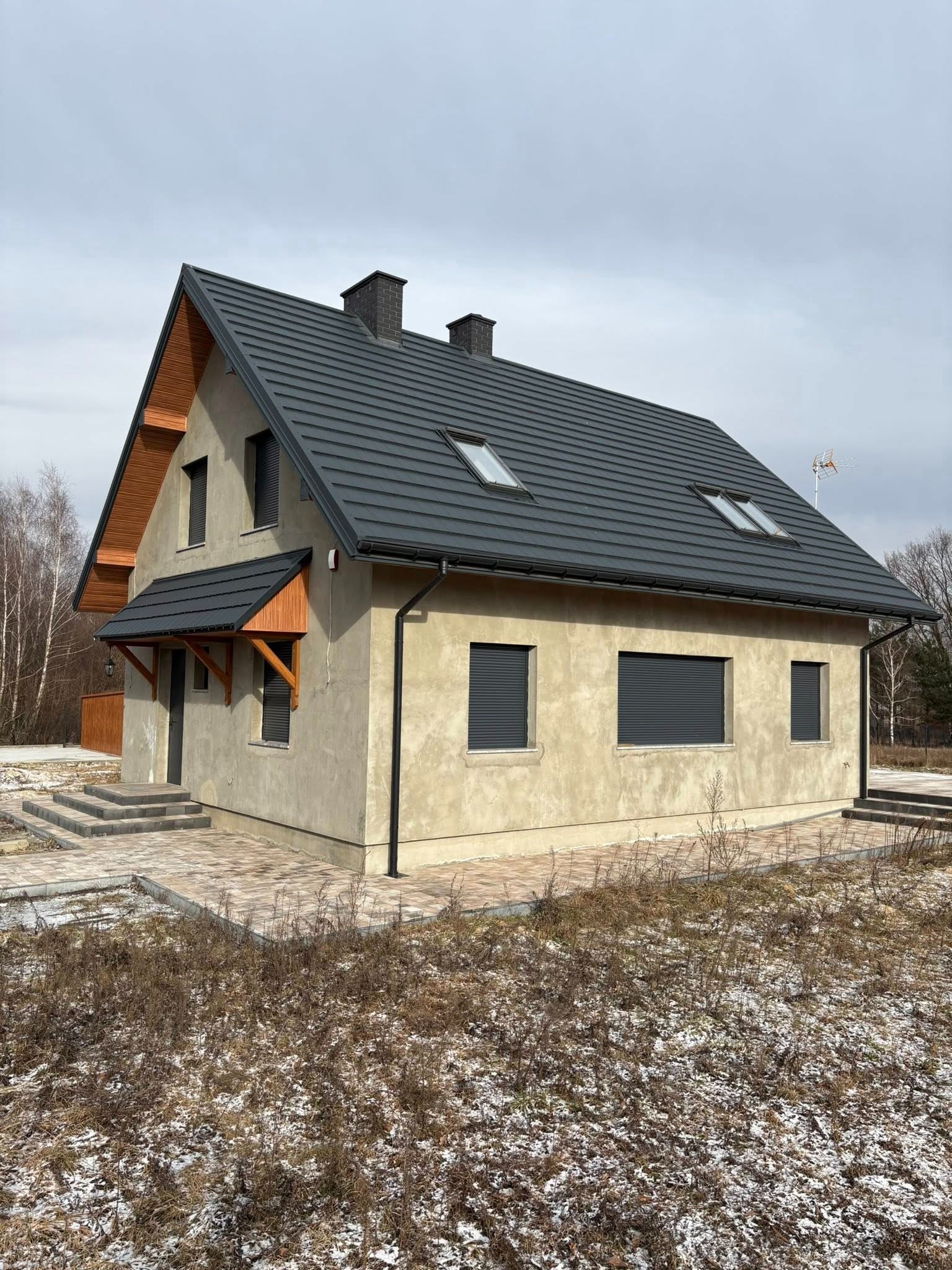 Nowoczesny dom jednorodzinny z szarą dachówką i beżową elewacją. Widoczne okna z roletami zewnętrznymi i drewniane elementy wykończeniowe nad wejściem.