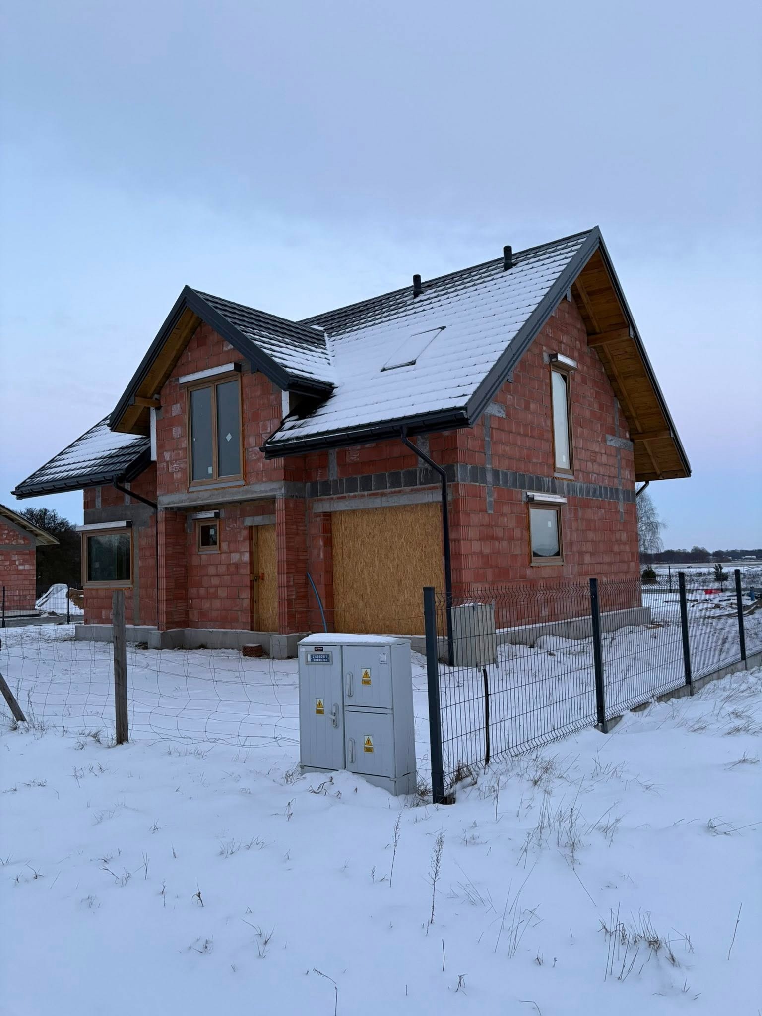 Niedokończony, dwukondygnacyjny dom z czerwonej cegły w zimowej scenerii. Widoczne okna z drewnianymi ramami i czarny dach pokryty śniegiem. Teren ogrodzony siatką.