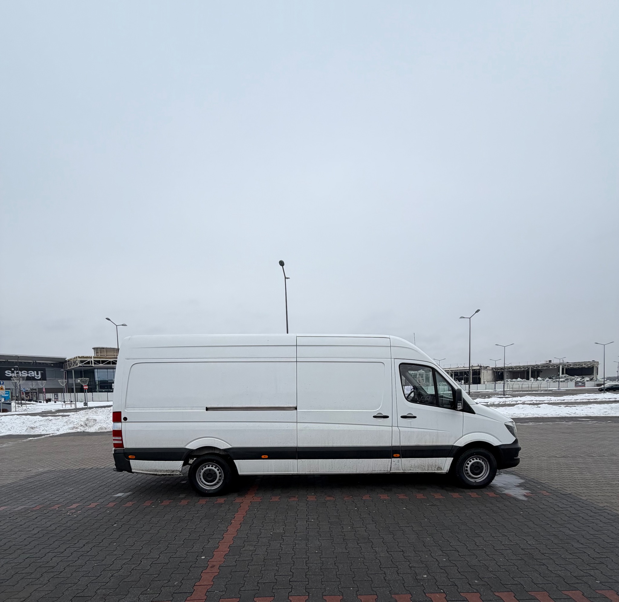 Biały, dostawczy van Mercedes Sprinter na parkingu z kostki, widok z boku, pochmurne niebo, w tle budynek sklepu Sinsay i resztki śniegu.