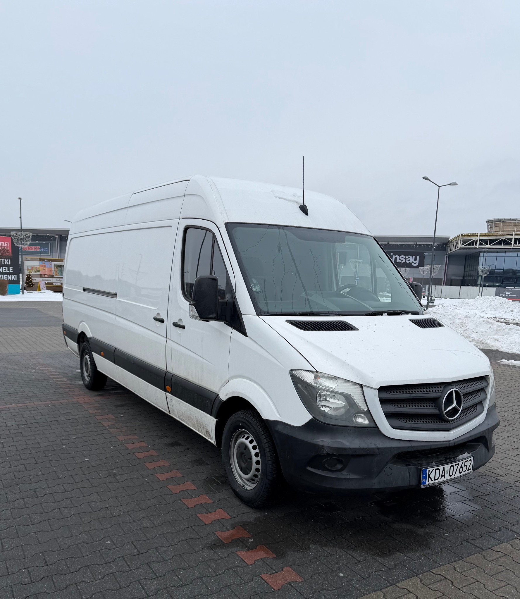 Biały, wysoki furgon Mercedes Sprinter na parkingu przed centrum handlowym w pochmurny dzień. Widoczna tablica rejestracyjna KDA 07652.