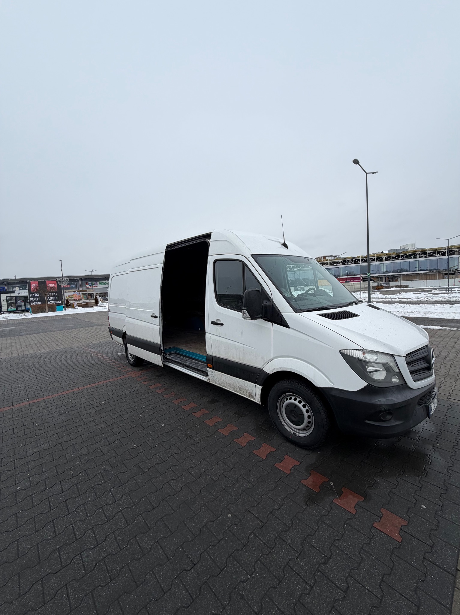 Biały bus Mercedes Sprinter z otwartymi drzwiami bocznymi na parkingu z kostki, w tle budynek handlowy i pochmurne niebo. Widok z boku, lekki kąt.