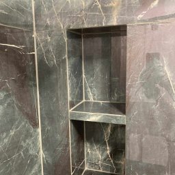 AM AUREUS WYKOŃCZENIA WNĘTRZ - Wykończona wnęka w kabinie prysznicowej, wyłożona płytkami imitującymi marmur w odcieniach szarości i bieli. Widoczne fugi i półka wewnątrz wnęki.
