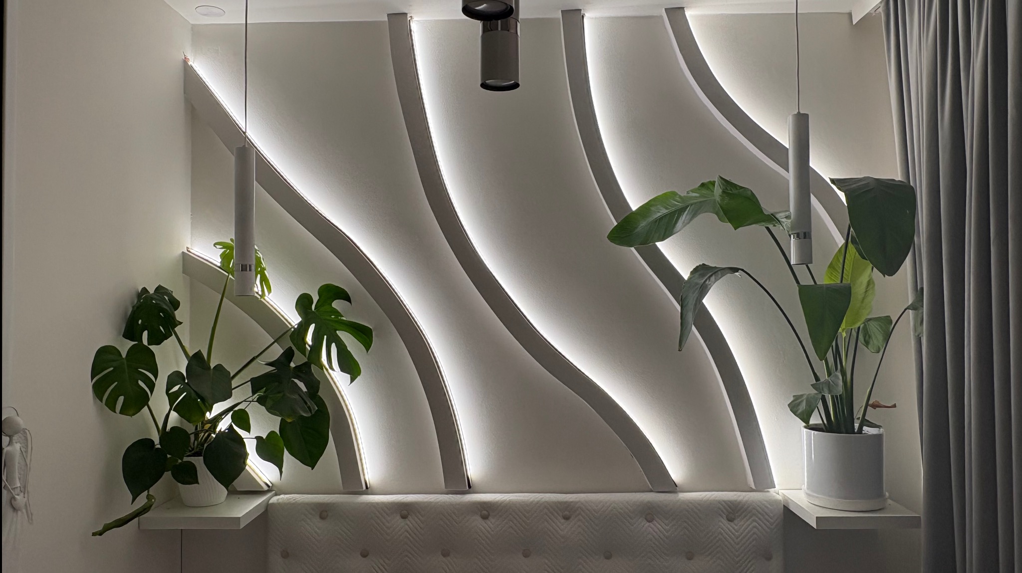 Biała ściana z dekoracyjnymi, podświetlanymi listwami, roślinami w doniczkach i lampami wiszącymi. Nowoczesny design wnętrza z subtelnym oświetleniem.