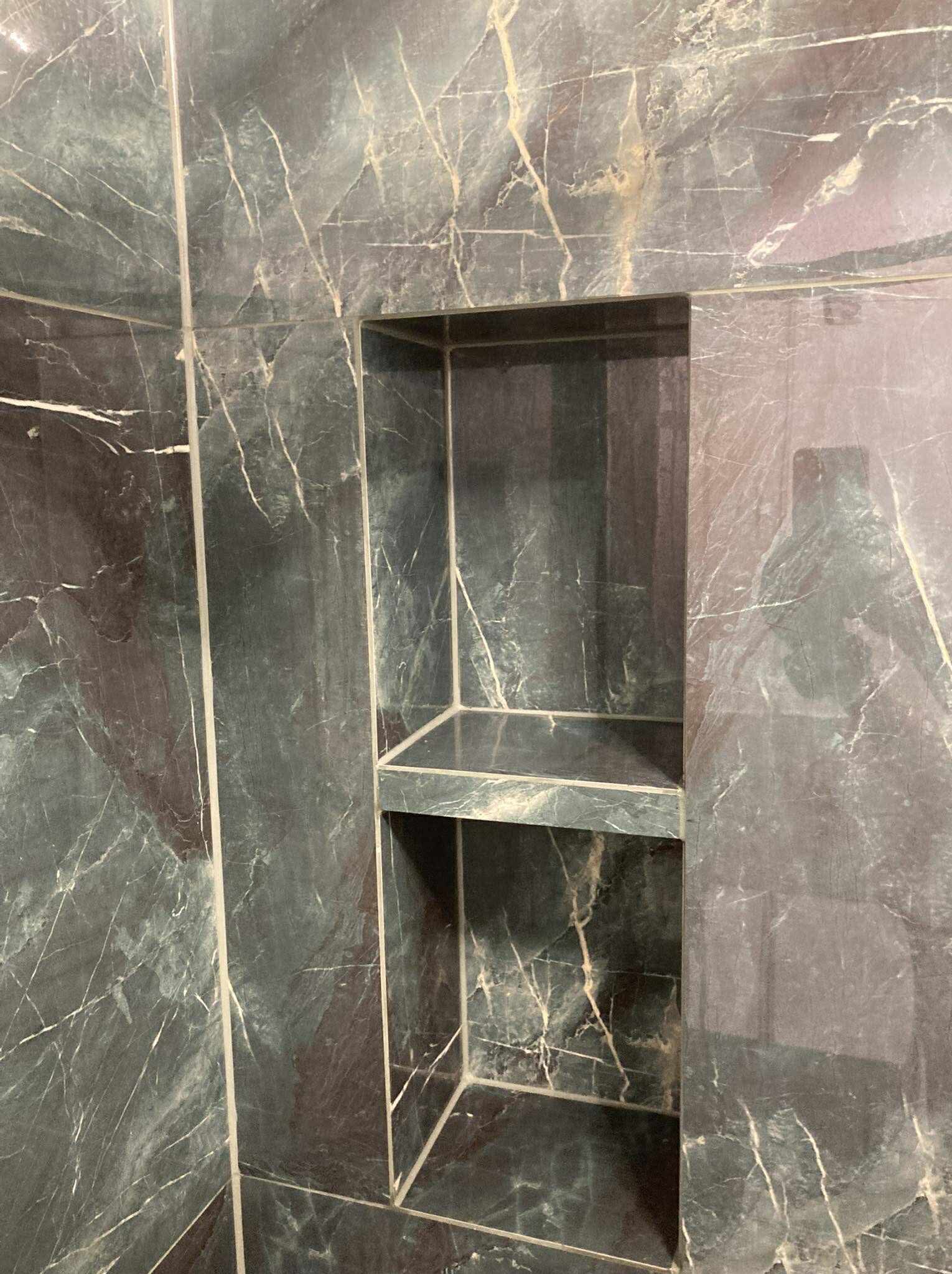 Wykończona wnęka w kabinie prysznicowej, wyłożona płytkami imitującymi marmur w odcieniach szarości i bieli. Widoczne fugi i półka wewnątrz wnęki.