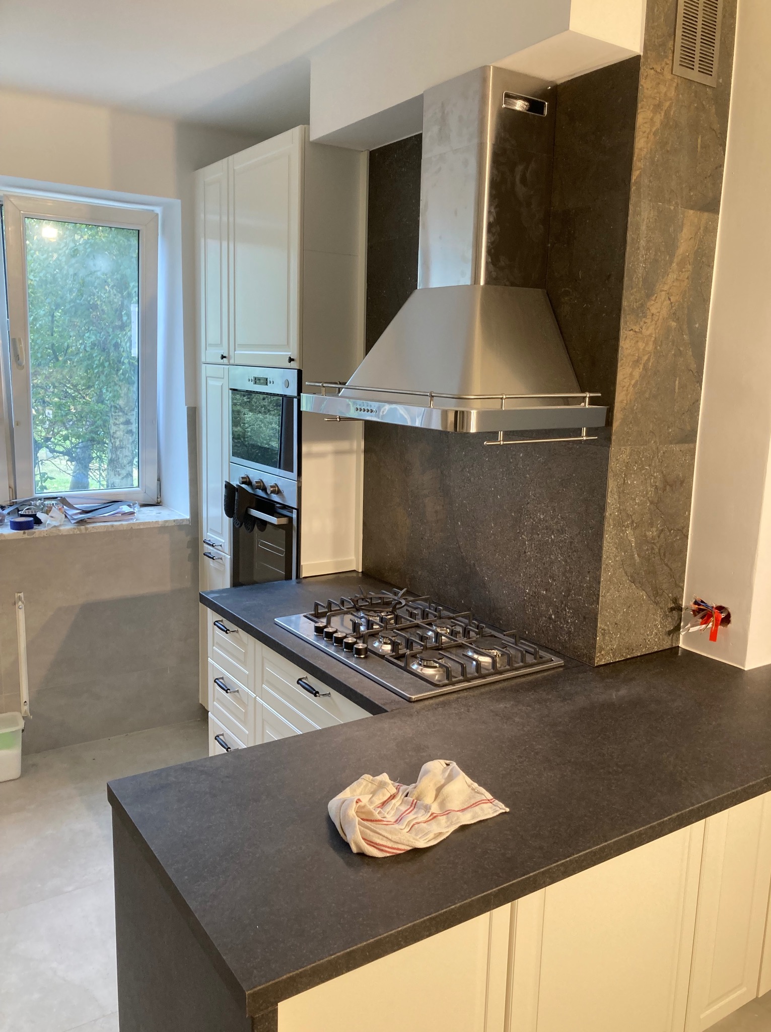 Kuchnia po remoncie: zabudowa z białymi szafkami, czarny blat, płyta gazowa i okap. Widoczne płytki imitujące kamień na ścianie i okno z widokiem na zieleń.