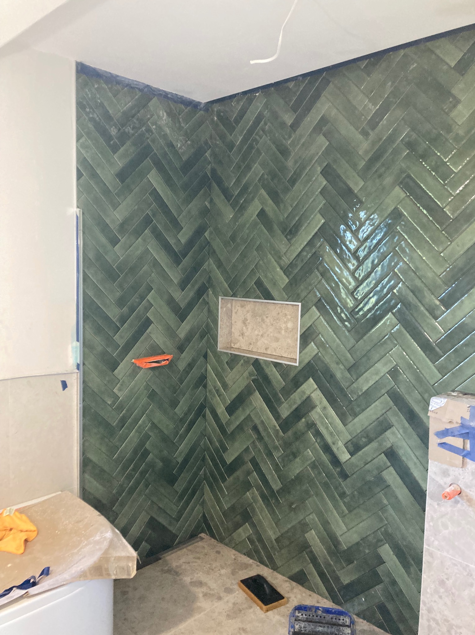 Ściana prysznica wyłożona zieloną, błyszczącą glazurą w jodełkę. Widoczna wnęka. Na podłodze narzędzia glazurnicze i resztki zaprawy. Taśma malarska zabezpiecza krawędzie.