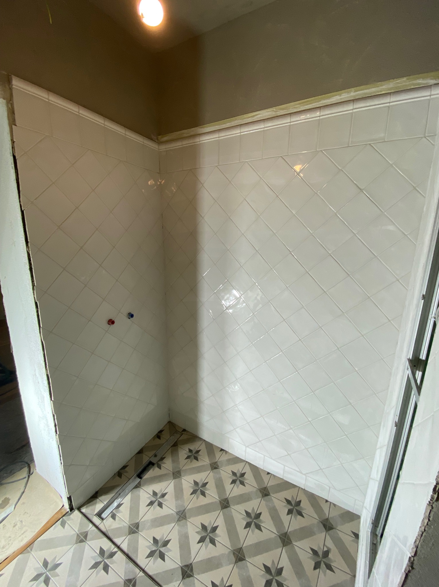 Wykończona kabina prysznicowa z białymi, ukośnymi płytkami na ścianach i wzorzystą podłogą. Widoczne przyłącza wodne i listwa wykończeniowa.