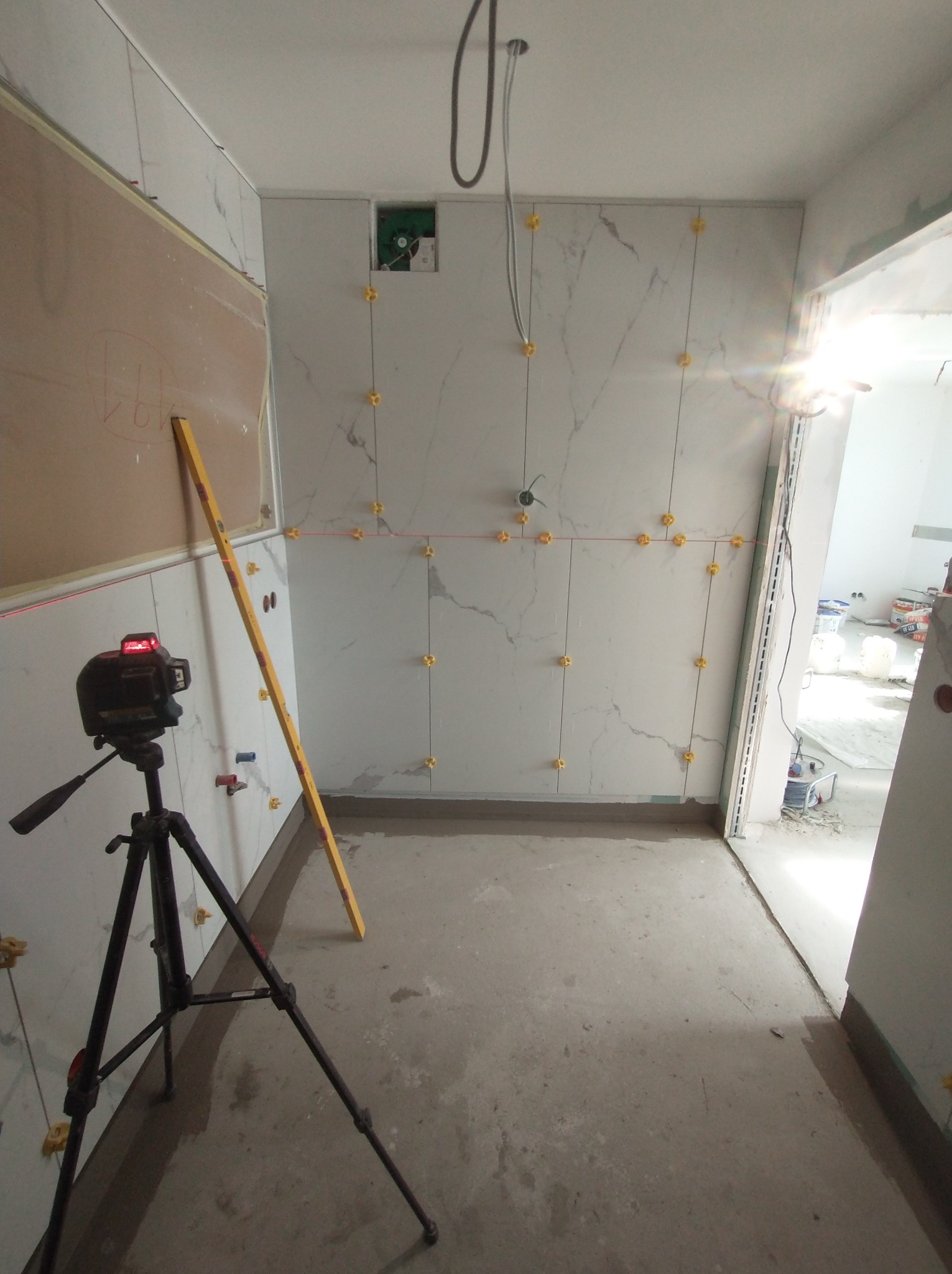 Układanie płytek marmurowych w łazience, widoczne poziomice laserowe i tradycyjne, żółte krzyżyki dystansowe, surowy beton na podłodze, etap prac wykończeniowych.