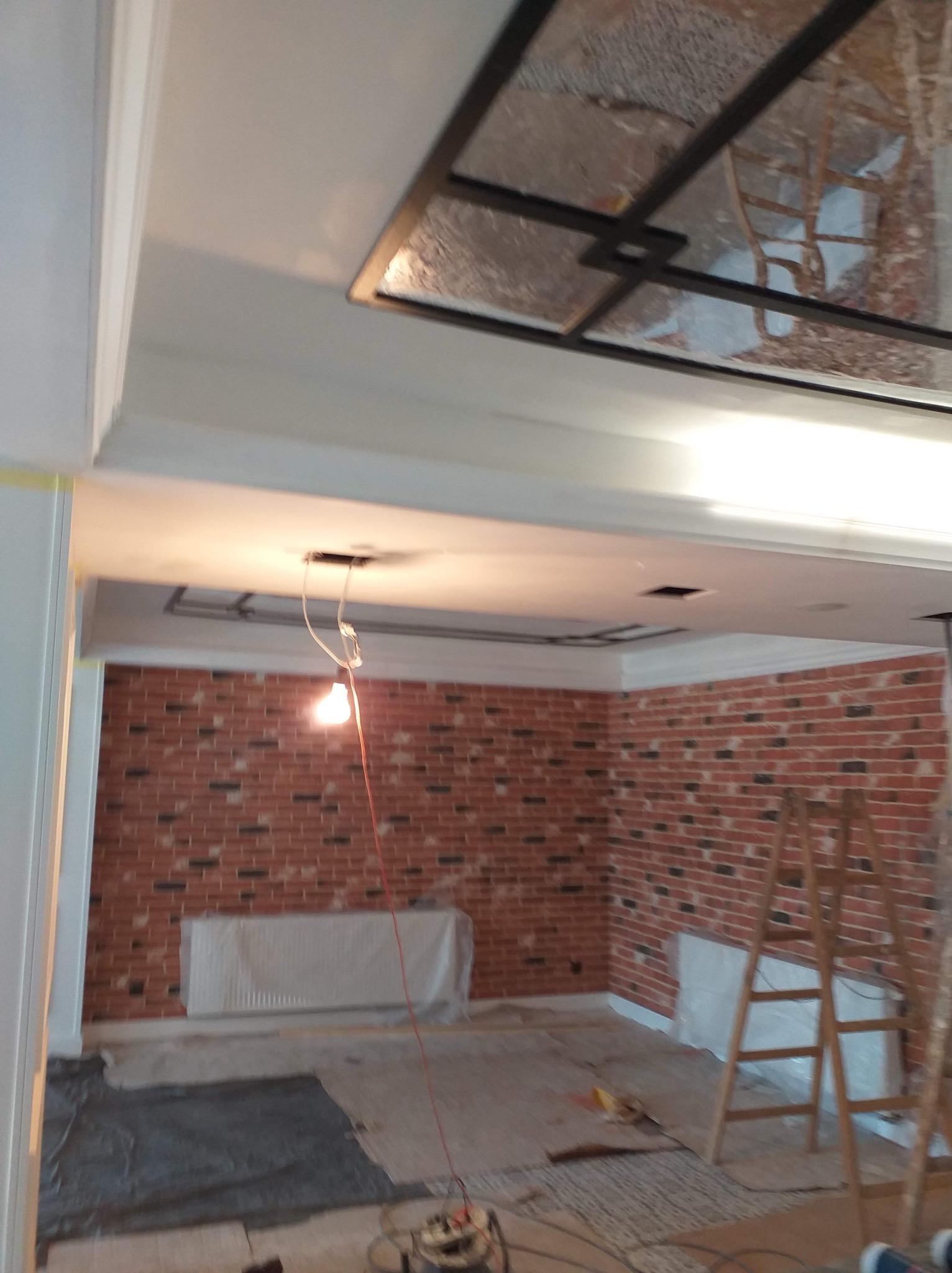Wnętrze w trakcie prac wykończeniowych: ceglana ściana, zabezpieczony grzejnik, drabina, widoczne kable i lampa wisząca. Prace remontowe w toku.