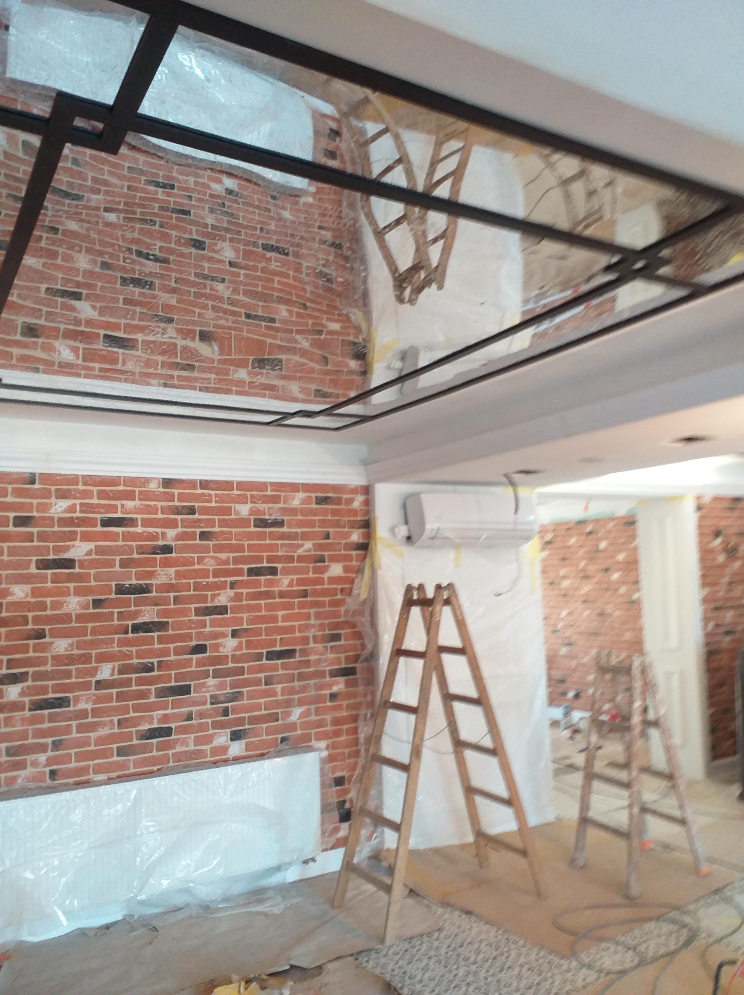 Wnętrze w trakcie prac wykończeniowych: ściana z cegły, drabiny malarskie, zabezpieczone folią elementy. Widoczna instalacja sufitowa i klimatyzator.