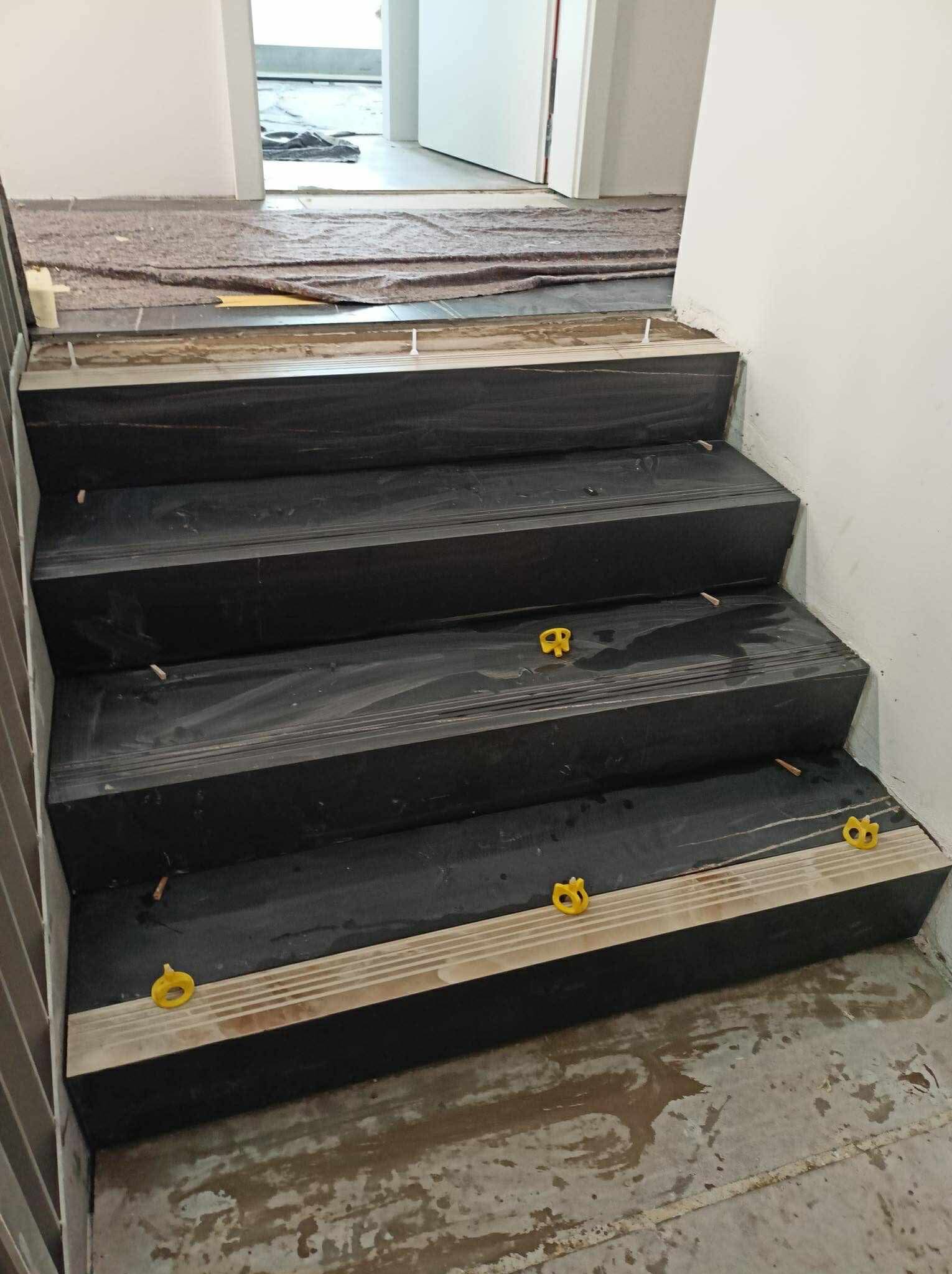 Montaż czarnych płytek na schodach z użyciem żółtych klinów dystansowych. Widoczne przygotowane podłoże i fragment zabezpieczonej podłogi w tle. Prace glazurnicze w trakcie.