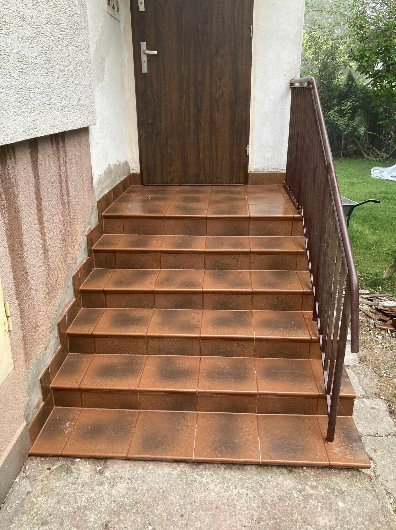 Wyłożone brązowymi płytkami schody wejściowe z metalową barierką. Widoczne drzwi wejściowe i fragment elewacji budynku. Tło stanowi zieleń ogrodu.