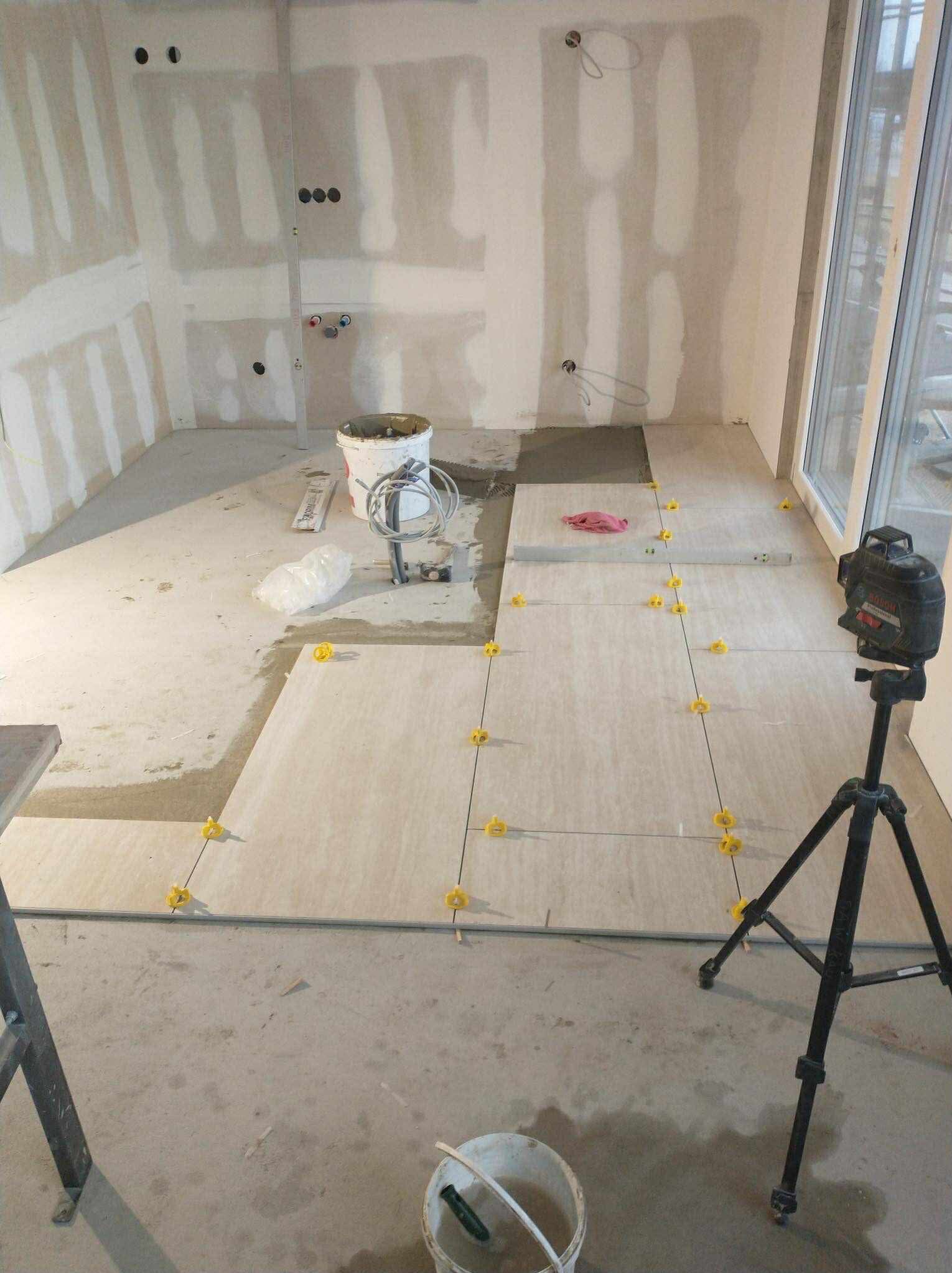 Układanie jasnych płytek podłogowych z użyciem poziomicy laserowej i krzyżyków dystansowych w surowym wnętrzu z przygotowanymi ścianami pod wykończenie.