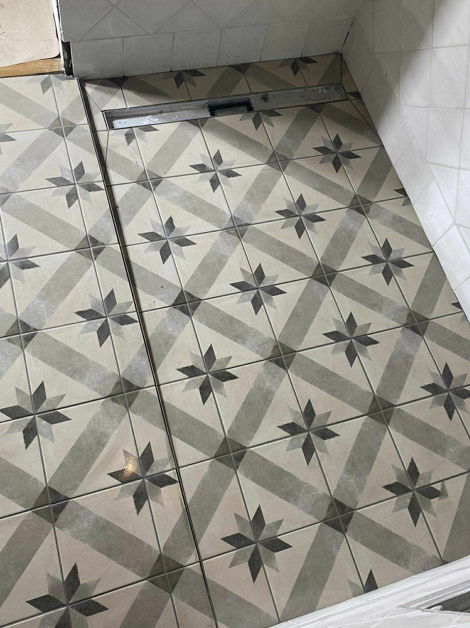 Ułożona podłoga w łazience z szarych płytek w geometryczny wzór gwiazdy i kraty, z widocznym odpływem liniowym. Górny kadr.