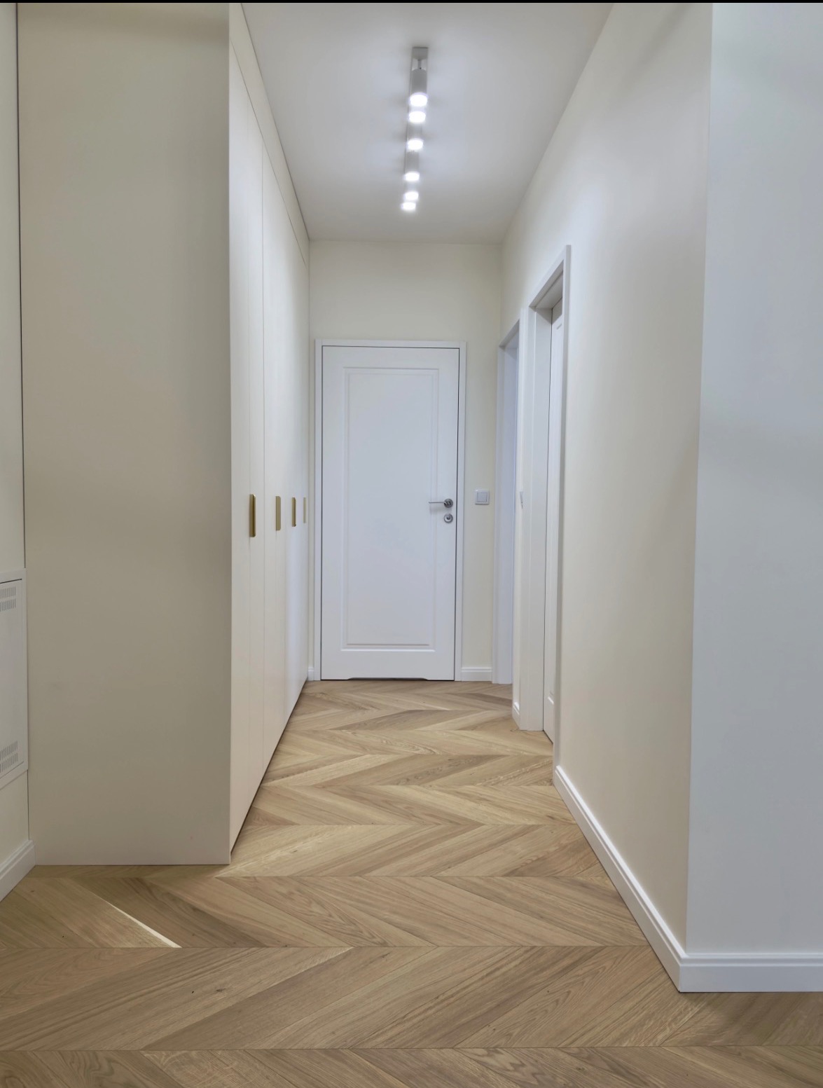 Jasny korytarz z parkietem w jodełkę, białe ściany i szafa w zabudowie z minimalistycznymi uchwytami. Oświetlenie sufitowe punktowe.