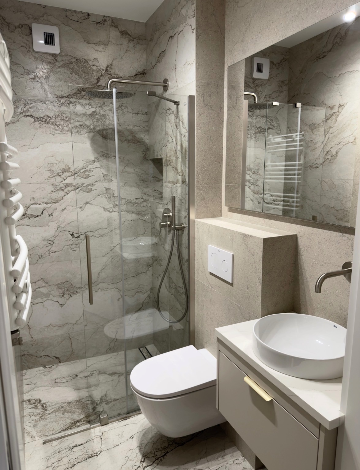 Wykończona łazienka z kabiną prysznicową typu walk-in, umywalką nablatową i podwieszaną toaletą. Płytki imitujące marmur na ścianach i podłodze. Nowoczesny design.