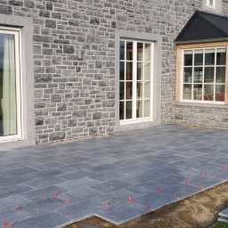 Piotr Wasiluk - Układanie szarej kostki brukowej na tarasie domu z kamienną elewacją. Widoczny mini-ładowarka i czerwone krzyżyki dystansowe. Częściowo ułożona nawierzchnia.