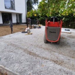 Piotr Wasiluk - Przygotowanie podłoża pod kostkę brukową: ubity tłuczeń, obrzeża betonowe, mini-wywrotka Thwaites. W tle budynek i pracownik. Widok na plac budowy w trakcie prac.