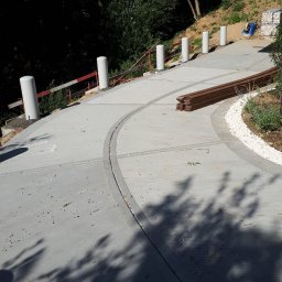 Piotr Wasiluk - Betonowa ścieżka z odpływem liniowym, obrzeżem z białego kamienia i drewnianymi elementami, prowadząca pod górę w otoczeniu zieleni.