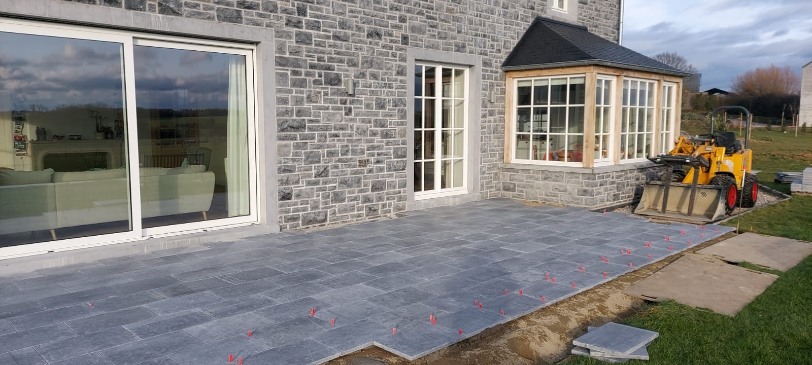 Układanie szarej kostki brukowej na tarasie domu z kamienną elewacją. Widoczny mini-ładowarka i czerwone krzyżyki dystansowe. Częściowo ułożona nawierzchnia.