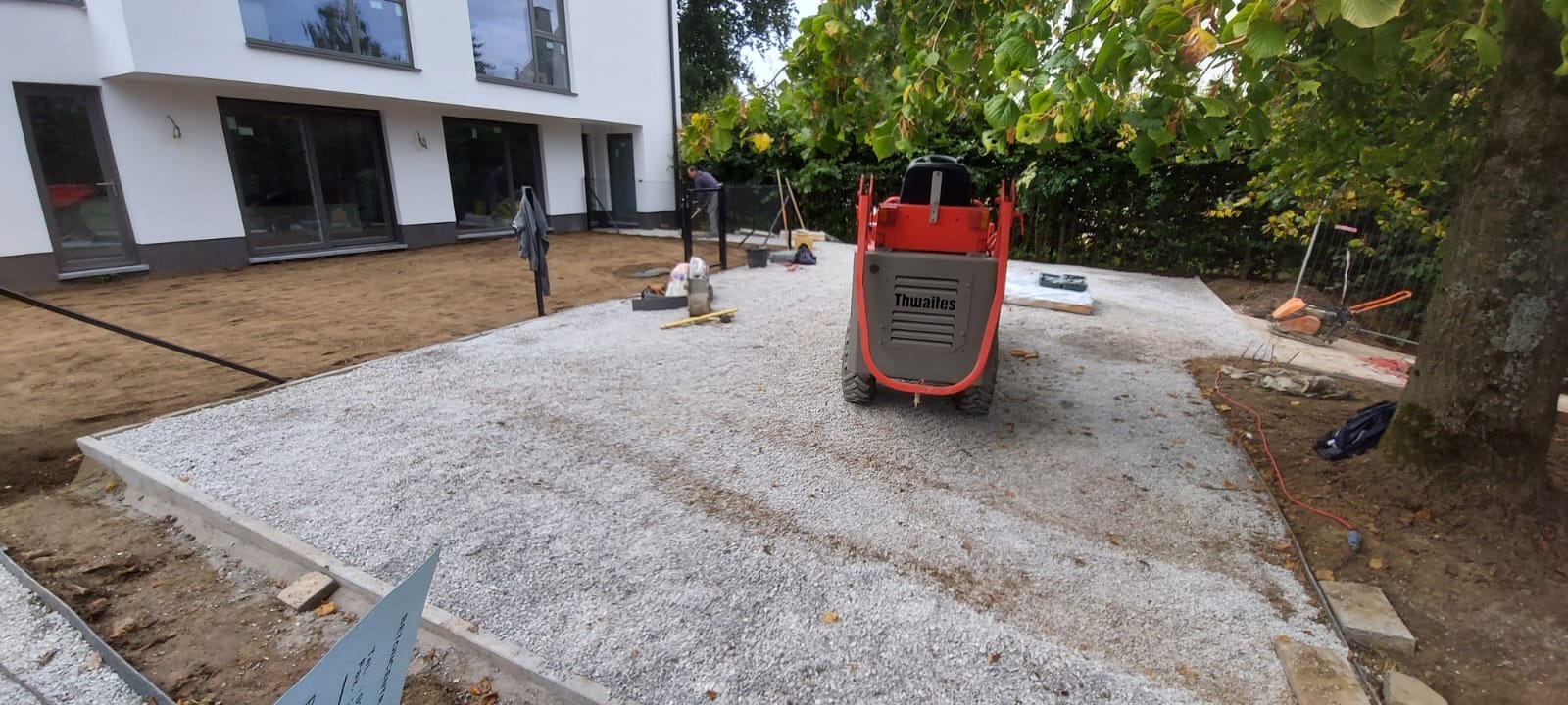 Przygotowanie podłoża pod kostkę brukową: ubity tłuczeń, obrzeża betonowe, mini-wywrotka Thwaites. W tle budynek i pracownik. Widok na plac budowy w trakcie prac.