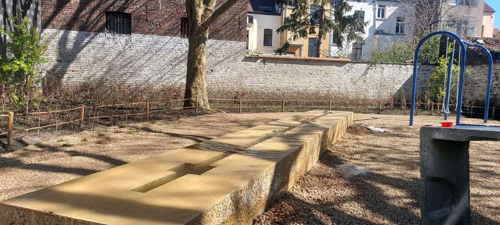 Ogród z elementami wodnymi: długa, kamienna fontanna z płynącą wodą, żwirowe podłoże, drewniane ogrodzenie, huśtawka w tle, ceglany mur. Słoneczny dzień.