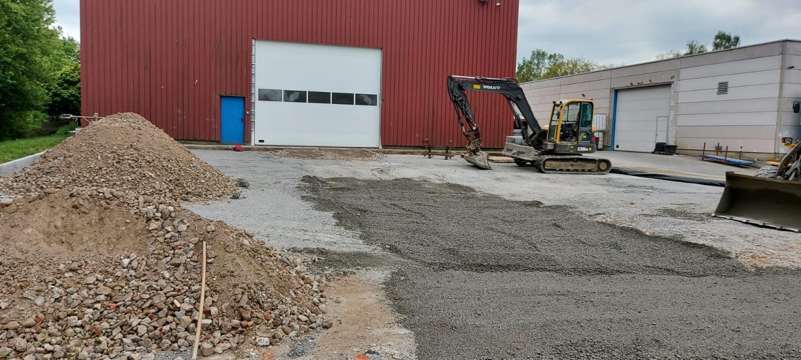 Żwirowa nawierzchnia wyrównana pod budowę, z koparką Volvo i stertą gruzu w tle, przed budynkiem z czerwoną ścianą i białą bramą garażową.