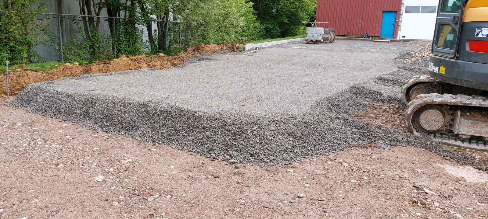Przygotowany teren pod kostkę brukową z użyciem kruszywa, widoczny fragment koparki i wytyczone granice. Etap prac przed ułożeniem kostki.