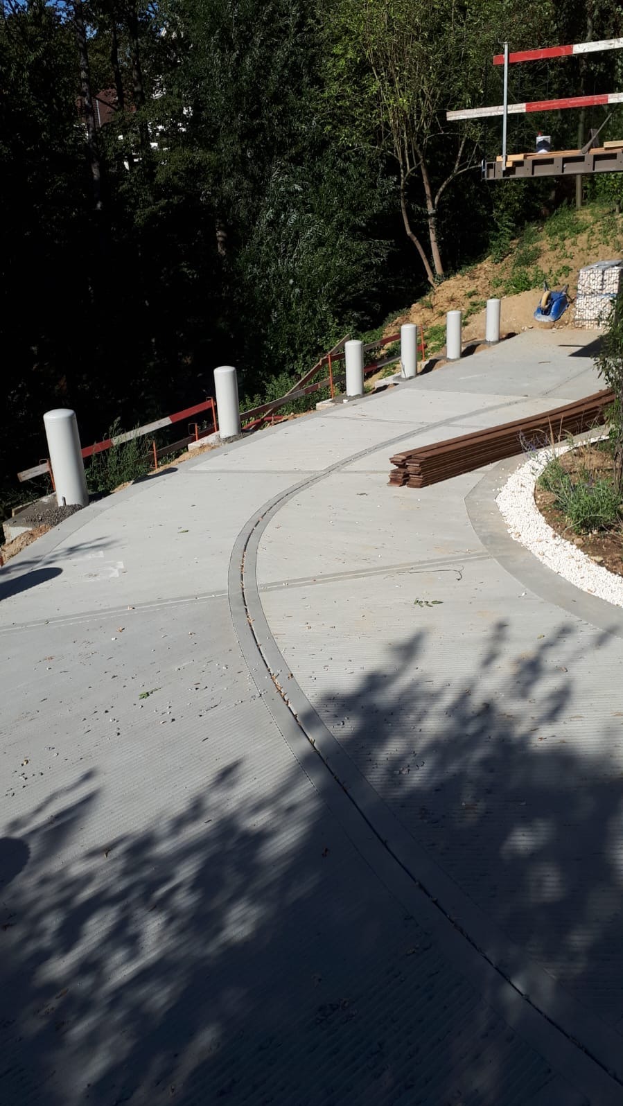 Betonowa ścieżka z odpływem liniowym, obrzeżem z białego kamienia i drewnianymi elementami, prowadząca pod górę w otoczeniu zieleni.