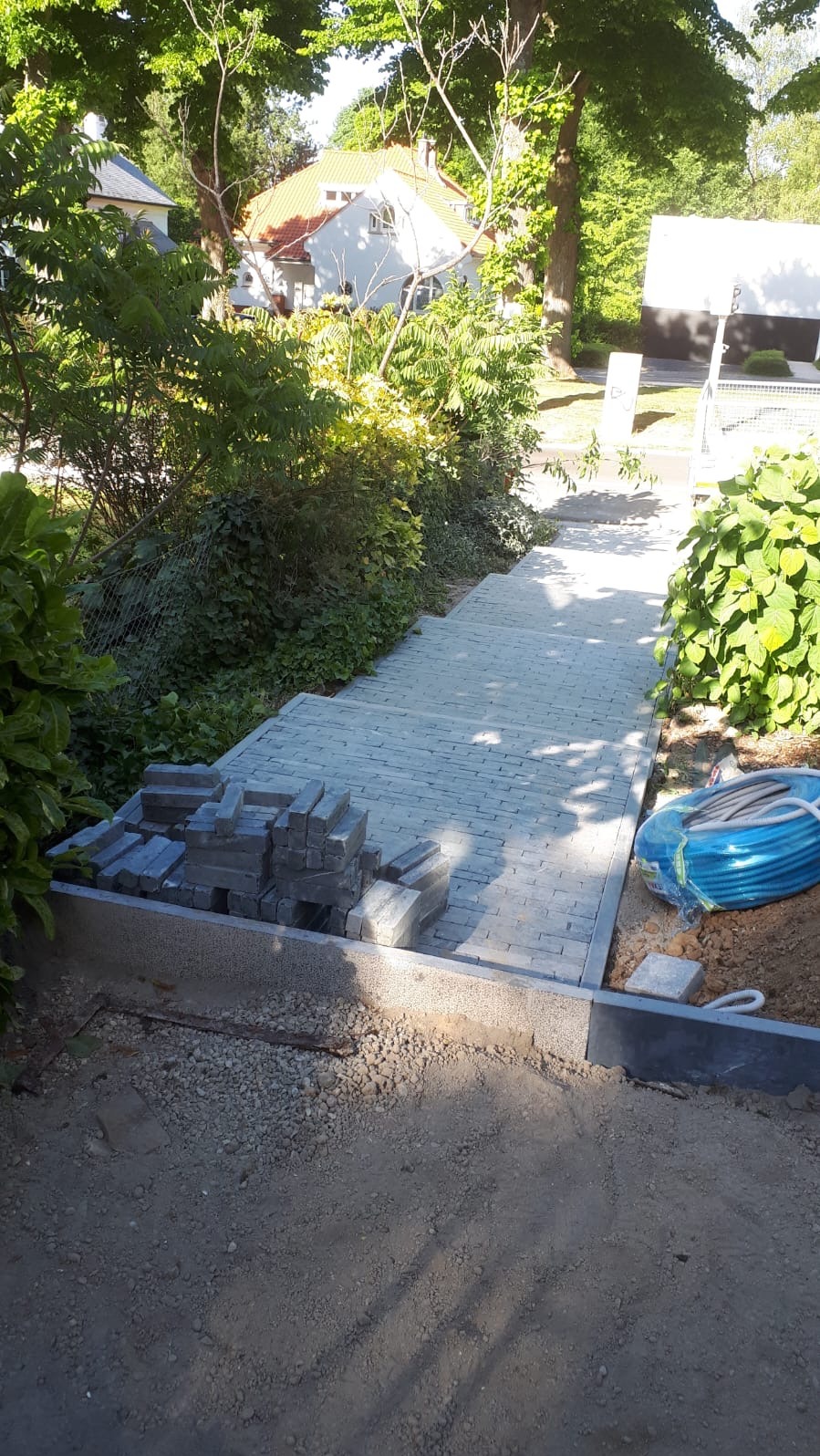 Schody z kostki brukowej w ogrodzie, częściowo ułożone, z widocznymi zapasami kostki i elementami instalacji, otoczone zielenią i domem w tle.