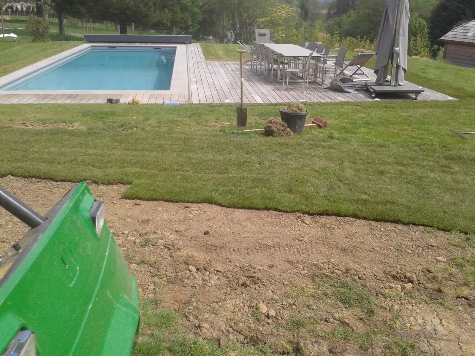 Fragment ogrodu z basenem w tle. Widoczny fragment trawy z rolki, narzędzia ogrodnicze i maszyna do układania trawy. W tle taras z meblami ogrodowymi.