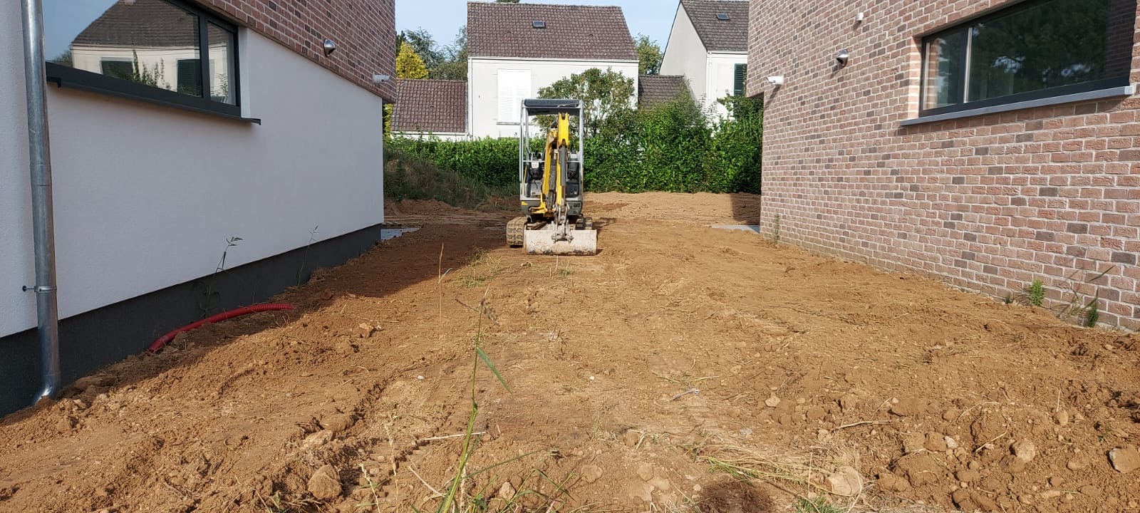 Żółta mini koparka na wyrównanym terenie budowy między dwoma budynkami z cegły i białym tynkiem. Przygotowanie pod trawnik lub kostkę brukową.