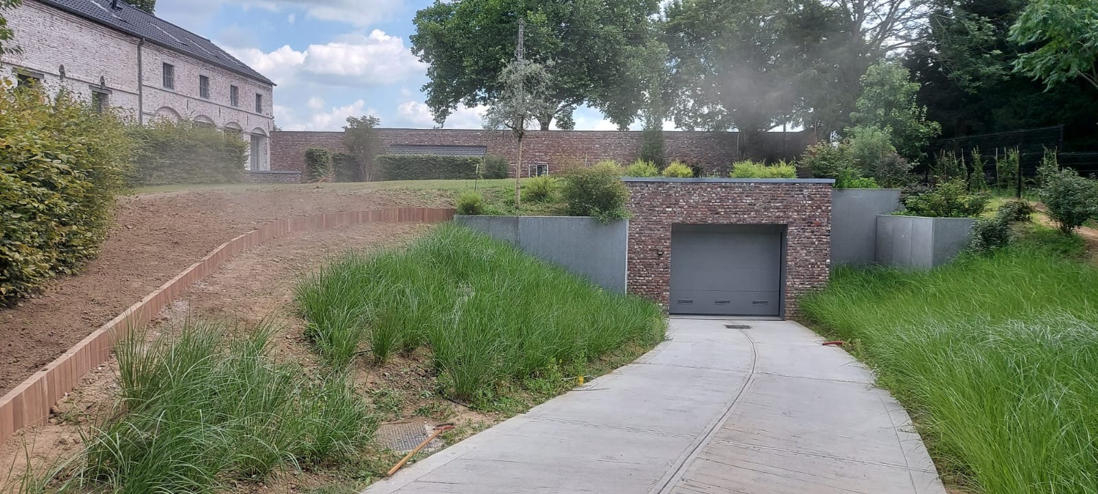 Nowoczesny ogród z podjazdem do garażu wkomponowanego w skarpę. Widoczne elementy architektury ogrodowej i przygotowany teren pod nasadzenia. Ujęcie w słoneczny dzień.