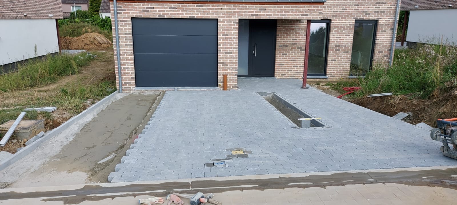 Układanie szarej kostki brukowej na podjeździe domu z cegły. Widoczny fragment korytka odwadniającego i narzędzia brukarskie. Częściowo wykonane obrzeże.