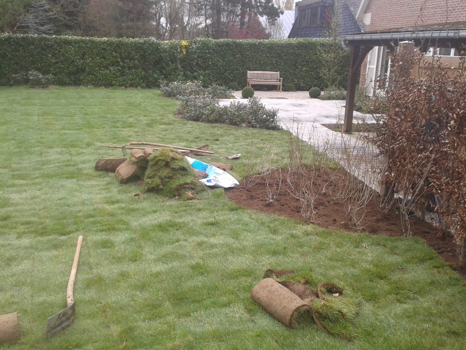 Fragment ogrodu w trakcie zakładania: rozłożona trawa z rolki, narzędzia ogrodnicze i świeżo posadzone krzewy na tle zielonego trawnika i żywopłotu.