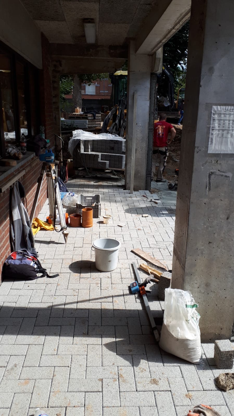 Układanie jasnoszarej kostki brukowej na zewnątrz budynku. Widoczne narzędzia, materiały budowlane i częściowo ułożona nawierzchnia. W tle koparka i pracownik.