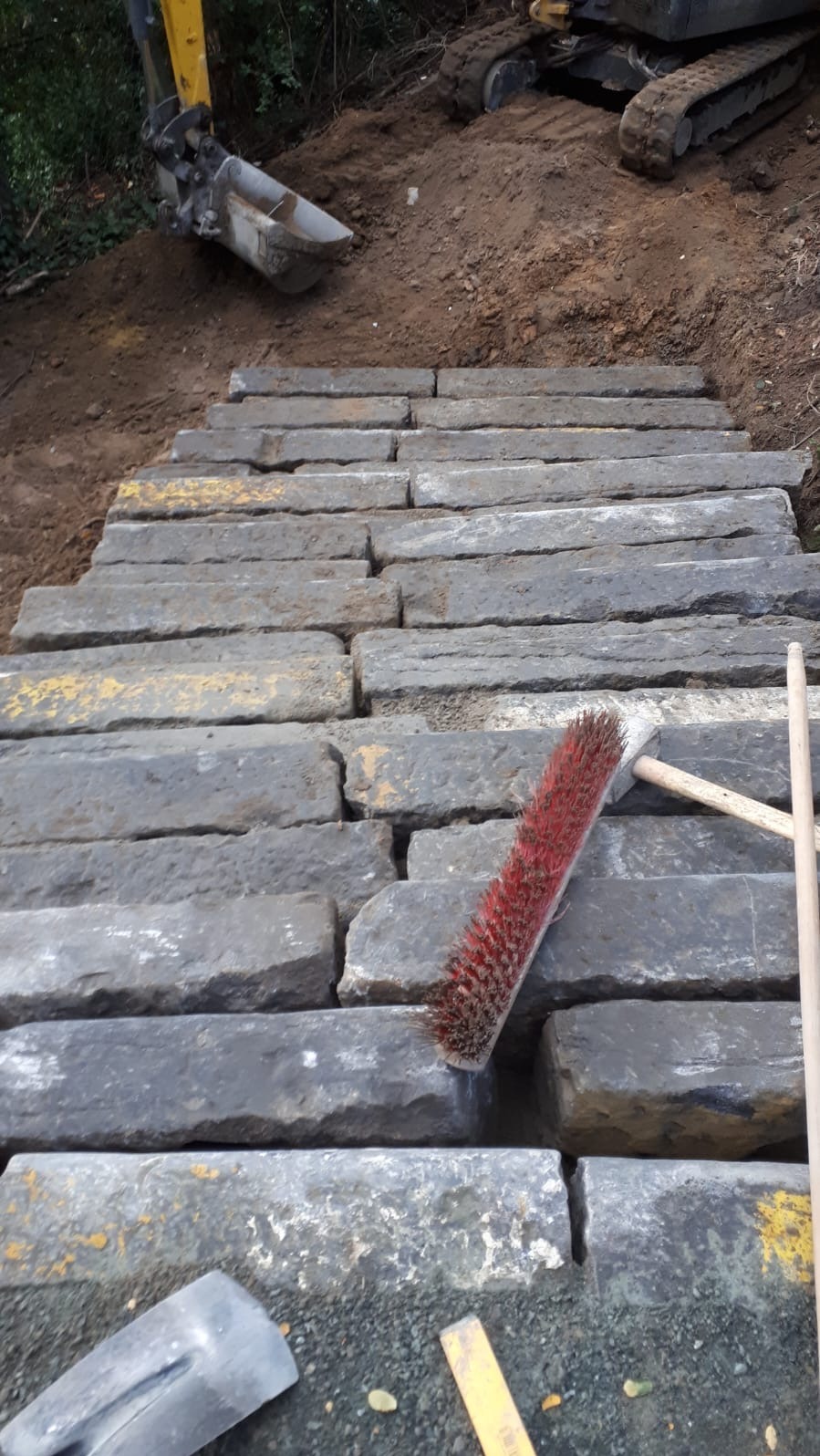 Kamienne stopnie schodów w trakcie budowy na zboczu, z widocznym sprzętem budowlanym i narzędziami, takimi jak szczotka i łopata, sugerujące prace brukarskie.